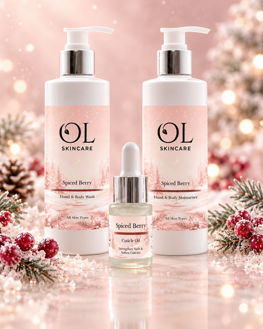 Three OL Skincare products arranged together on a reflective surface: two large pump bottles labeled Spiced Berry Hand & Body Wash and one smaller dropper bottle labeled cuticle Oil, set against a soft pink holiday-themed background with pine branches and red berries.