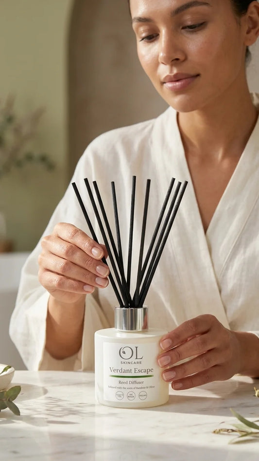 A woman in a white robe holds a reed diffuser bottle labeled 'OL Scents Verdant Escape' with black reeds, set on a light marble surface in a softly lit indoor setting.