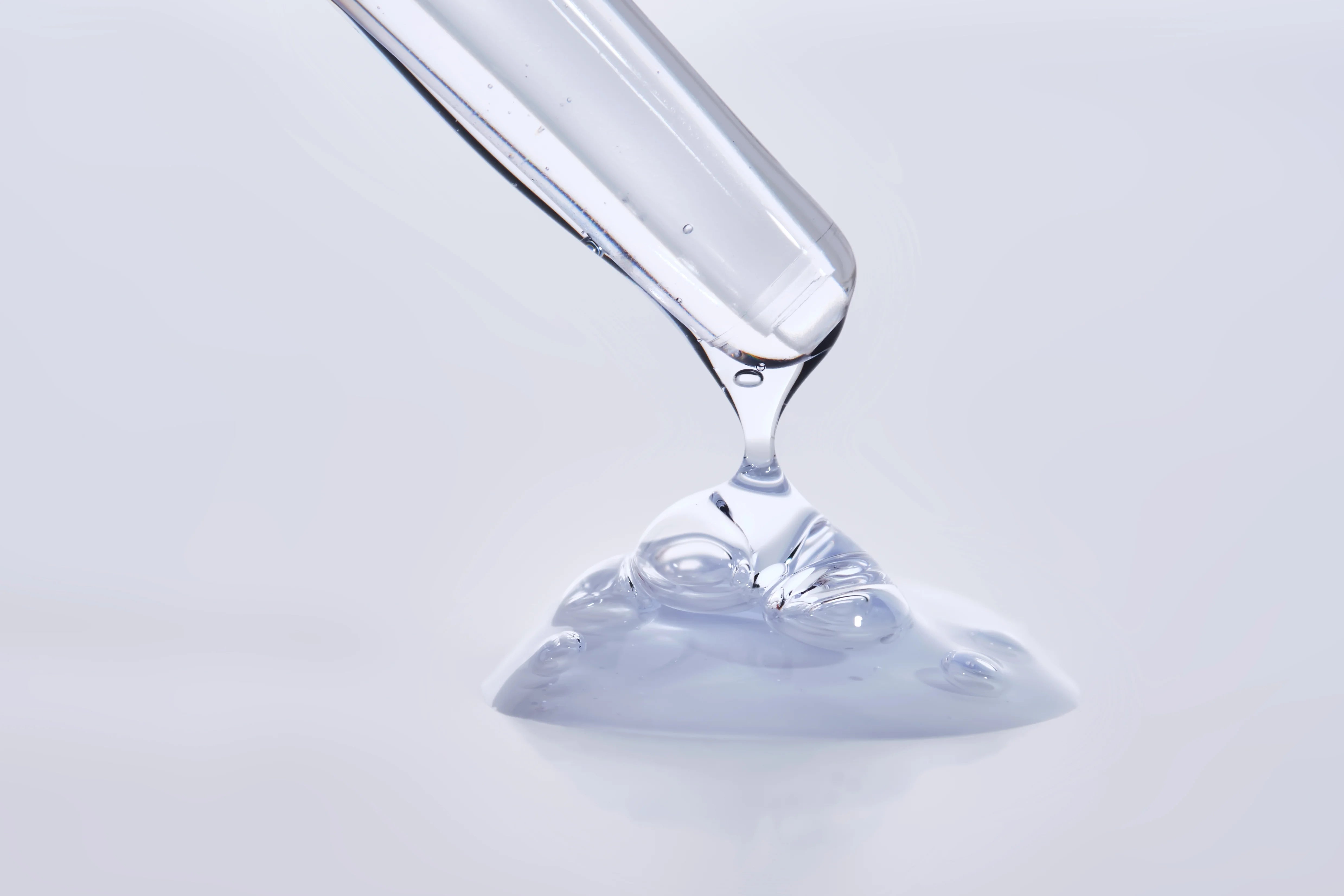 Clear liquid glycerine being poured from a glass container onto a light gray background