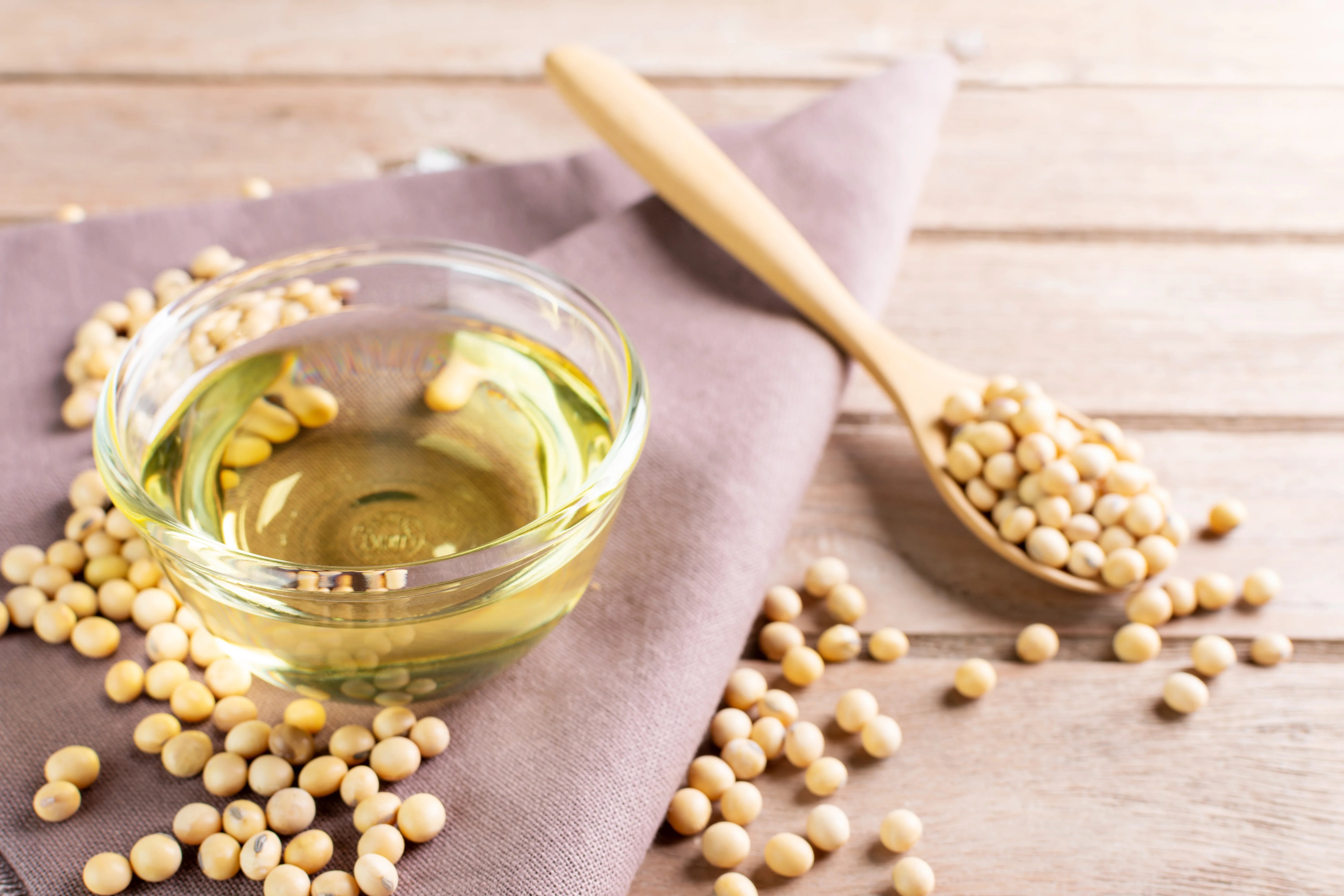 Glass bowl of soybean oil with scattered soybeans on a wooden surface