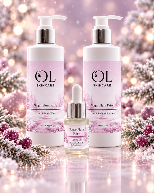 Three OL Skincare products in a coordinated holiday-themed setting: two large pump bottles labeled Sugar Plum Fairy Hand & Body Wash and one smaller dropper bottle labeled cuticle Oil, arranged with pink floral and snowy decorative elements on a reflective surface.
