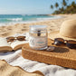 A jar of OL Skincare SPF 50 Derm-Veil sunscreen sits on a woven tray on a beach blanket, surrounded by a straw hat, sunglasses, and an open book with the ocean and palm trees in the background.