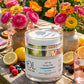 A clear cosmetic jar labeled 'OL Skincare Derma Veil SPF 50' sits on a wooden table outdoors, surrounded by flowers, lemons, berries, and gold-toned cutlery in a bright garden setting.