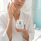 A woman in a white robe applies cream to her face while holding a white cosmetic jar with a silver lid in a bright bathroom setting.