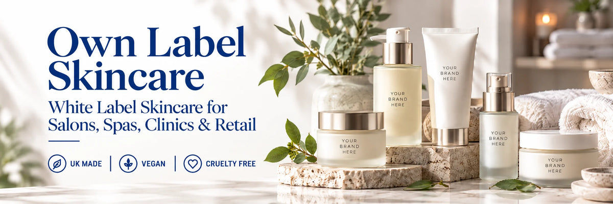 A promotional banner for 'Own Label Skincare' featuring several white and cream skincare bottles and jars with gold caps arranged on stone pedestals. The background depicts a softly blurred spa-like bathroom scene with a bathtub, folded towels, and green leaves.