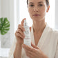 A woman in a white robe holds a white skincare pump bottle with a silver cap in front of a bright window. The bottle has a minimalist black logo and text that appears to read 'OL'.