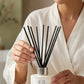 A woman in a white robe holds a reed diffuser bottle labeled 'OL Scents Verdant Escape' with black reeds, set on a light marble surface in a softly lit indoor setting.