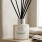 A reed diffuser bottle with black reed sticks sits on a stone ledge beside folded towels. The bottle is cream-colored with a silver cap and a label reading 'OL Skincare Verdam Escape Reed Diffuser'.