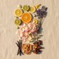 A styled flat-lay arrangement of dried botanical and citrus ingredients on a beige fabric background, including sliced oranges and limes, lavender sprigs, pale pink petals, small white flowers, vanilla beans, cinnamon sticks, and dark brown bark pieces.