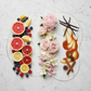 A styled flat-lay of a perfume or fragrance oil labeled 'Scent of Summer' on a white marble background. The oil is presented in an organic oval glass container, surrounded by decorative ingredients that evoke a warm summer scent profile: pink and white flowers in the center, sliced grapefruit, lemons, blueberries, vanilla beans, caramel sauce, cinnamon sticks, and star anise.